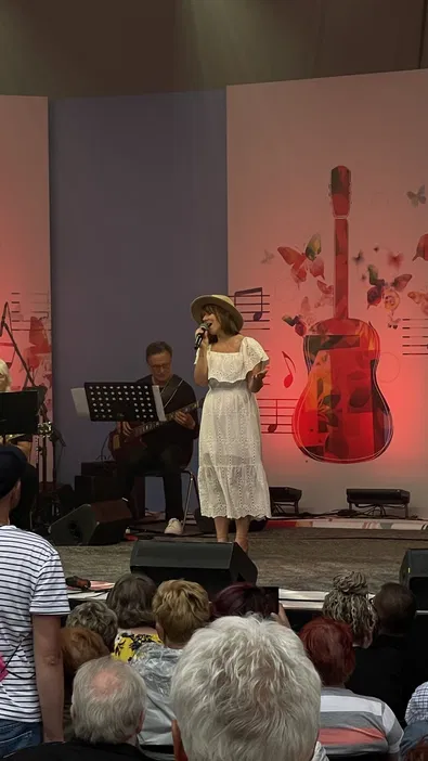 Koncert na żywo, śpiewaczka w białej sukience i kapeluszu na scenie z zespołem, sztuka gitarowa i tłum publiczności.