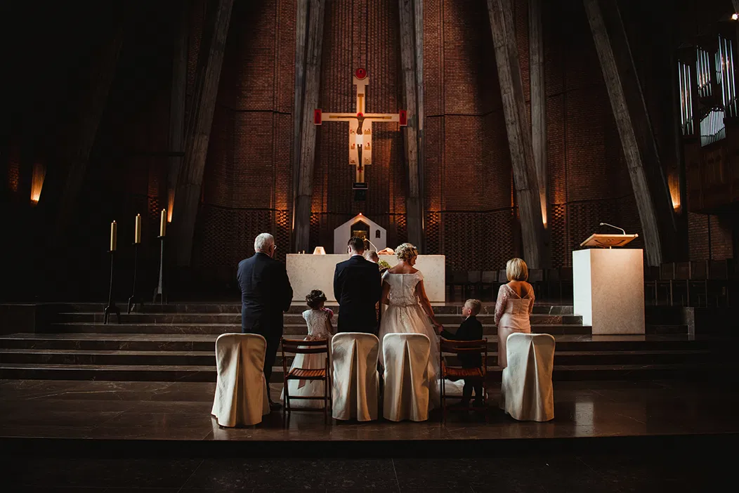 Wnętrze kościoła podczas ceremonii ślubnej, para młoda i goście przed ołtarzem, subtelne oświetlenie, atmosfera sakralna.