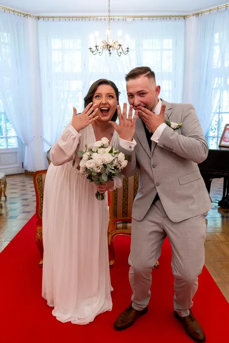 Para młoda pokazująca obrączki na ceremonii ślubnej, elegancko ubrana, z bukietem białych kwiatów na czerwonym dywanie.