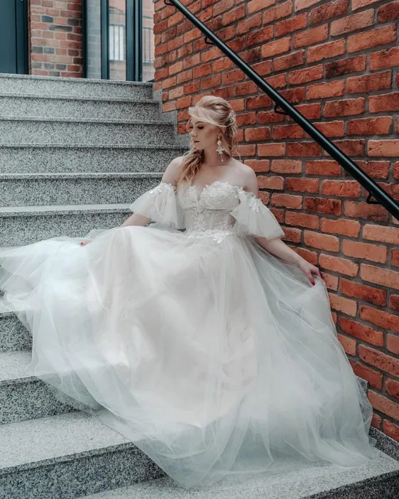 Kobieta w eleganckiej sukni ślubnej siedzi na schodach z ceglaną ścianą w tle.
