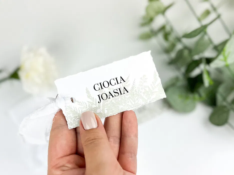 Ręka trzyma kartkę z napisem Gioia Joasia, tło z zielonymi liśćmi i białym kwiatem.