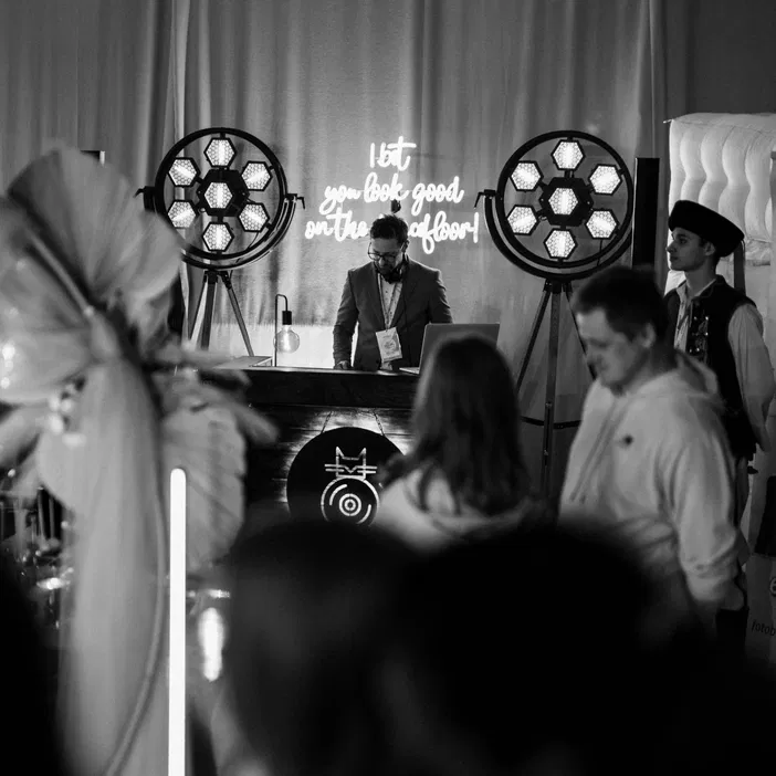 Czarno-białe zdjęcie z DJ-em na imprezie, w tle neonowe napisy i dekoracje świetlne.