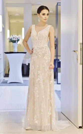  Kobieta w eleganckiej, koronkowej, kremowej sukni wieczorowej stojąca w nowoczesnym wnętrzu. 