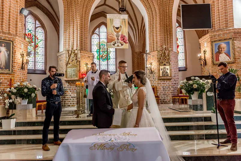  Ślub w kościele katolickim, para młoda przed ołtarzem podczas ceremonii z księdzem, dekoracje kwiatowe i witraże w tle. 