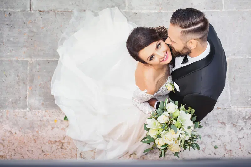  Para młoda przytula się na tle kamiennej posadzki, panna młoda w białej sukni trzyma bukiet białych kwiatów, romantyczna fotografia ślubna z lotu ptaka. 
