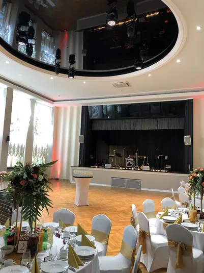  Elegancka sala bankietowa z ustawionymi stołami, ozdobami kwiatowymi i sceną przygotowaną na przyjęcie weselne lub event. 