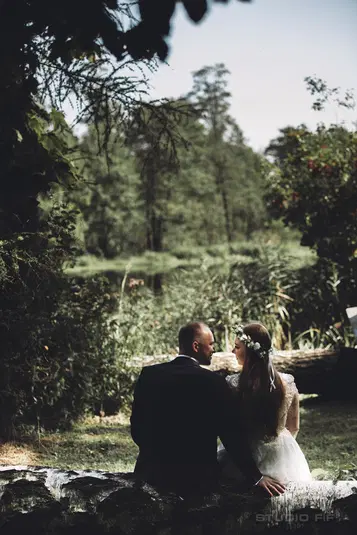  Para młoda siedząca nad jeziorem w lesie, romantyczna sesja ślubna na świeżym powietrzu, panna młoda w wianku, pan młody w garniturze, naturalna sceneria, fotografia ślubna w plenerze. 