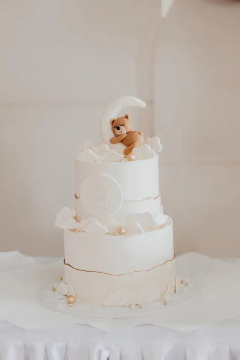 Biały tort piętrowy z dekoracją w postaci misia śpiącego na księżycu, ozdobiony chmurkami i perłami, idealny na chrzest lub baby shower.