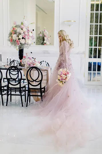  Elegancka panna młoda w różowej sukni ślubnej, trzymająca bukiet kwiatów, przy ozdobnym stole w jasnym wnętrzu z kwiatowymi dekoracjami i czarnymi krzesłami. 