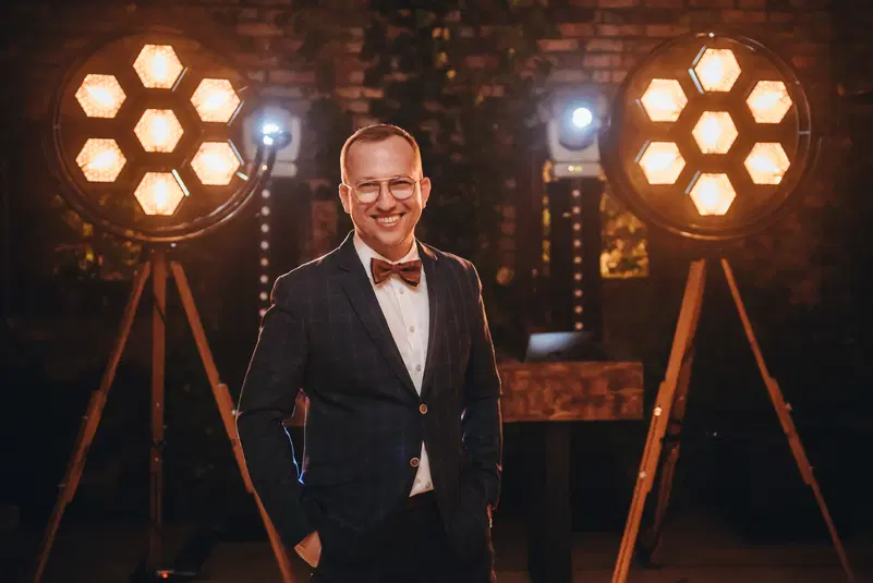  Mężczyzna w eleganckim garniturze i muszce stojący przed dekoracyjnymi lampami w industrialnym wnętrzu. 