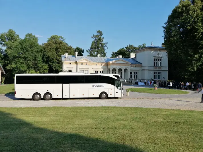  Autokar turystyczny przed elegancką posiadłością otoczoną zielenią podczas słonecznego dnia. 