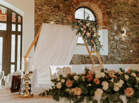 Dekoracja weselna z kamienną ścianą, kwiatami i drewnianą konstrukcją w eleganckim wnętrzu.