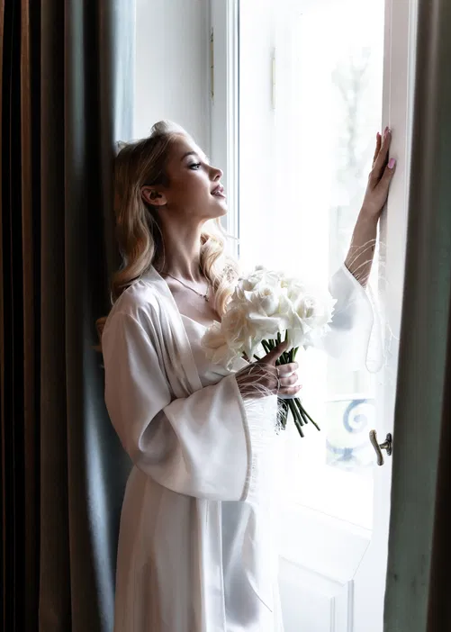 Blondynka w białym szlafroku przy oknie trzyma bukiet białych róż.