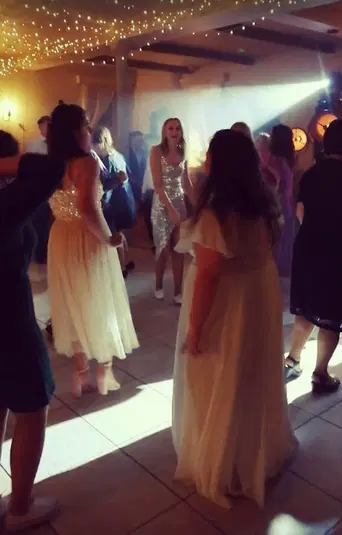  Grupa osób w eleganckich strojach tańczy na weselu pod ozdobnymi światłami i girlandami świetlnymi w przytulnym wnętrzu sali balowej. 