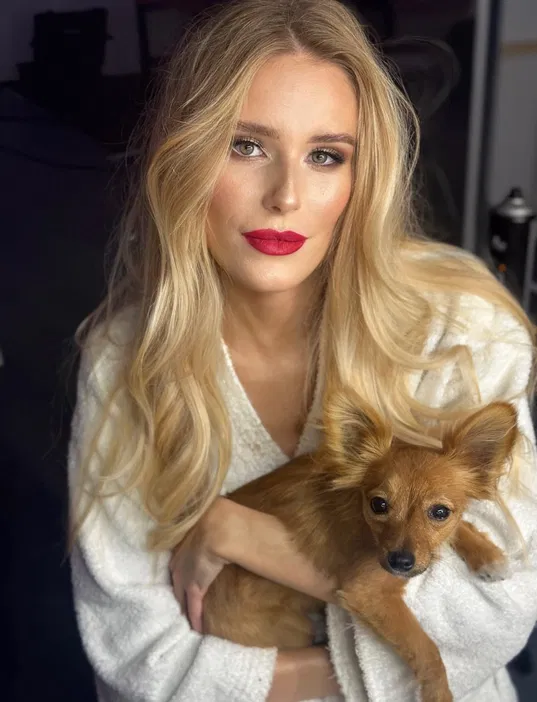 Blondynka z długimi włosami trzymająca małego brązowego psa w objęciach, uśmiechnięta, w białym szlafroku.
