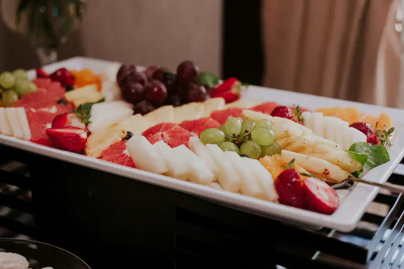  Taca z różnorodnymi owocami, w tym winogronami, truskawkami, melonem, ananasem i grejpfrutem, podana na eleganckim stole. 