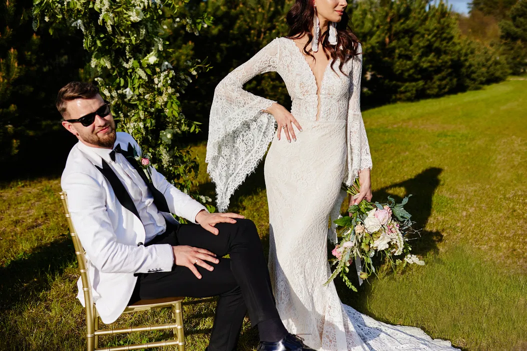 Para młoda podczas sesji ślubnej na świeżym powietrzu, mężczyzna w eleganckim garniturze, kobieta w białej koronkowej sukni i z bukietem kwiatów, zielone tło przyrody.