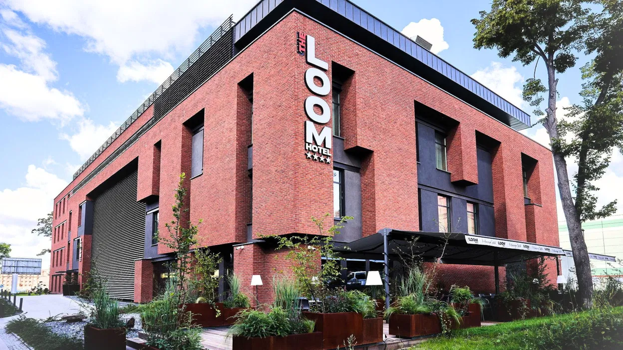 Czerwona fasada hotelu z widocznym logo „The Loom”, nowoczesny design, zielony krajobraz z drzewami i roślinnością na tle błękitnego nieba.