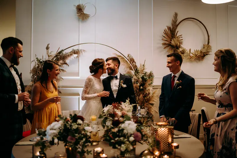 Para młoda świętująca przyjęcie weselne w eleganckim wnętrzu z dekoracjami z suszonych kwiatów i świecami, otoczona gośćmi, w atmosferze radości i przyjaźni. 