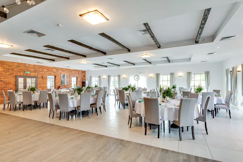  Elegancka sala bankietowa z okrągłymi stołami i krzesłami, dekoracje z zielonych roślin, ceglane ściany, jasne oświetlenie, przestrzeń na przyjęcia i wydarzenia. 