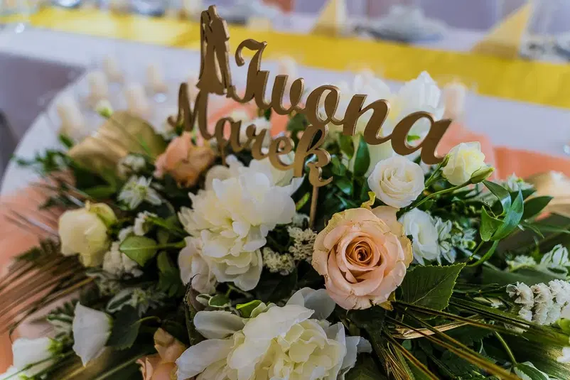  Dekoracja ślubna z kwiatami w odcieniach bieli i różu oraz personalizowanym napisem Iwona i Marek na weselnym stole. 