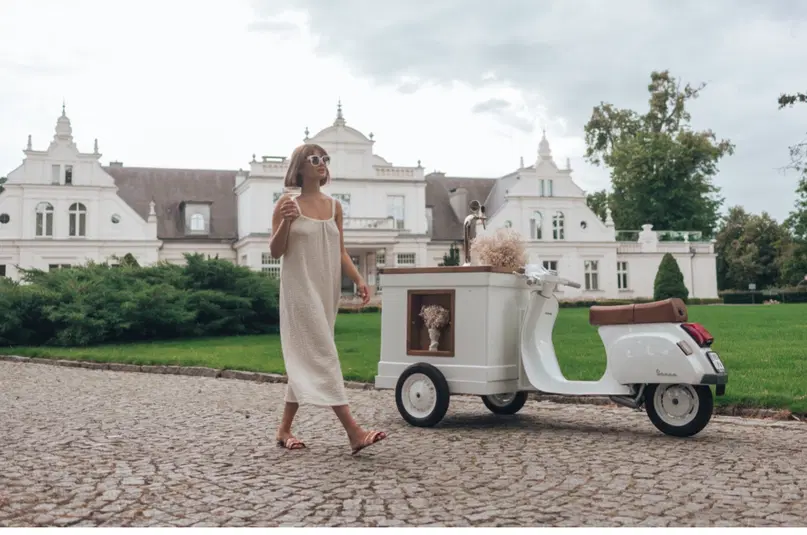  Kobieta w jasnej sukience spacerująca przy eleganckiej rezydencji z białym skuterem na tle zielonego trawnika. 