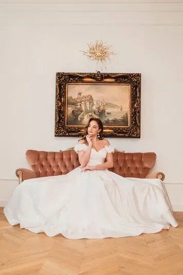  Kobieta w eleganckiej białej sukni siedzi na różowej sofie przed ozdobnym obrazem, w eleganckim wnętrzu. 