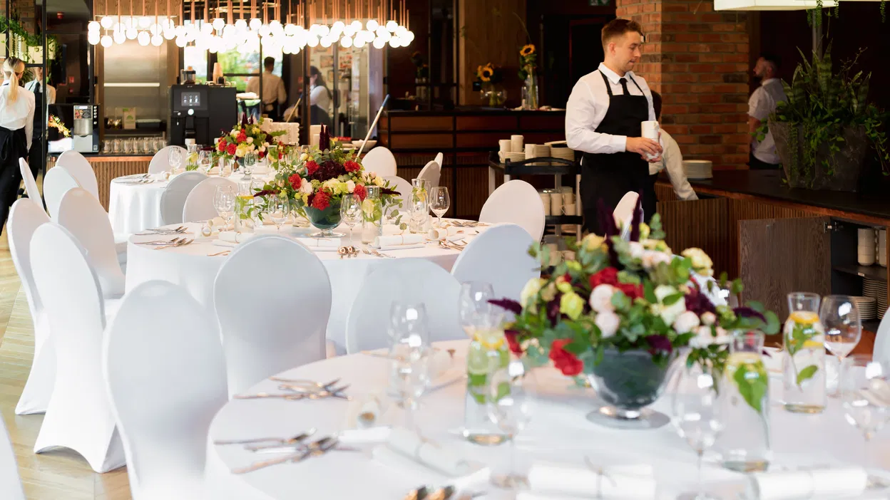 Elegancka sala bankietowa z dekoracjami kwiatowymi na białych obrusach, przygotowana na przyjęcie weselne.
