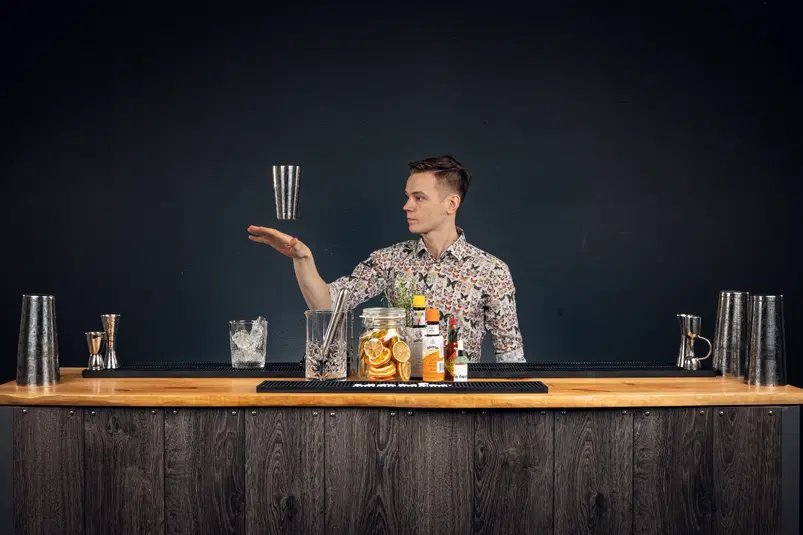  Barman w stylowej koszuli pokazuje sztuczki barmańskie, żonglując shakerem za barem wyposażonym w przybory do koktajli i składniki w słoikach oraz butelki alkoholi. 
