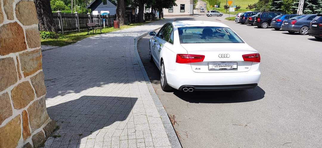  Biały Audi A6 TDI zaparkowany na chodniku obok brukowanego muru, w tle drzewa i zaparkowane samochody. 