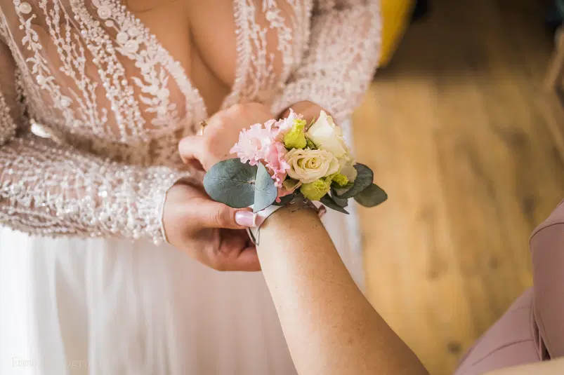  Panna młoda w koronkowej sukni przypinająca kwiatową bransoletkę na rękę druhny, elegancki bukiet z róż i eukaliptusa, stylowy ślub. 