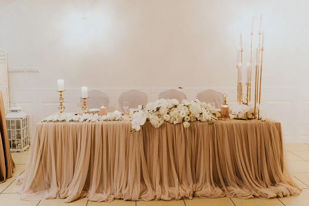 Elegancka dekoracja stołu weselnego z beżowym obrusem i białymi kwiatami, złote świeczniki i wysokie świece, romantyczna aranżacja ślubna, styl glamour.