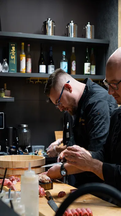 Kucharze przygotowujący dania w kuchni japońskiej, otoczeni butelkami sake i składnikami na sushi.