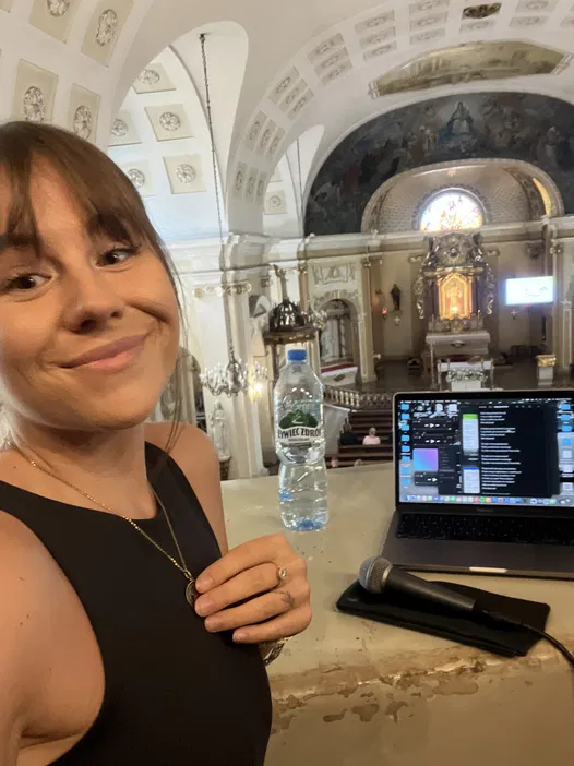 Kobieta z uśmiechem w kościele, w tle laptop, mikrofon i dekoracje sakralne.