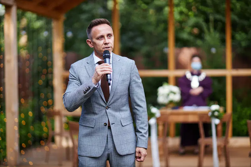  Mężczyzna w garniturze przemawiający do mikrofonu podczas ceremonii na świeżym powietrzu. 