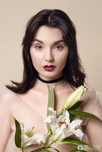  Kobieta z ciemnymi włosami i czerwoną szminką, trzymająca białe lilie na tle beżowej ściany, elegancki portret. 