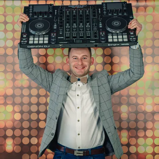  Mężczyzna w marynarce i muszce trzyma konsolę DJ uśmiechając się w świetle dyskotekowych świateł. 