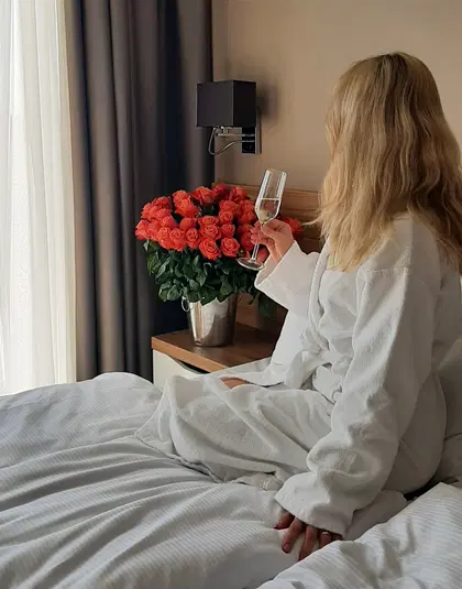  Blondynka w białym szlafroku siedzi na łóżku, trzymając kieliszek szampana, obok bukiet czerwonych róż w wazonie, elegancki pokój hotelowy. 