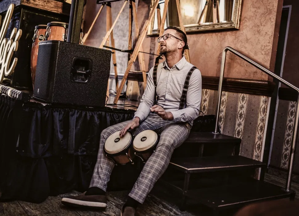 Mężczyzna grający na bongo siedzący na schodach przy scenie z instrumentami perkusyjnymi w tle.