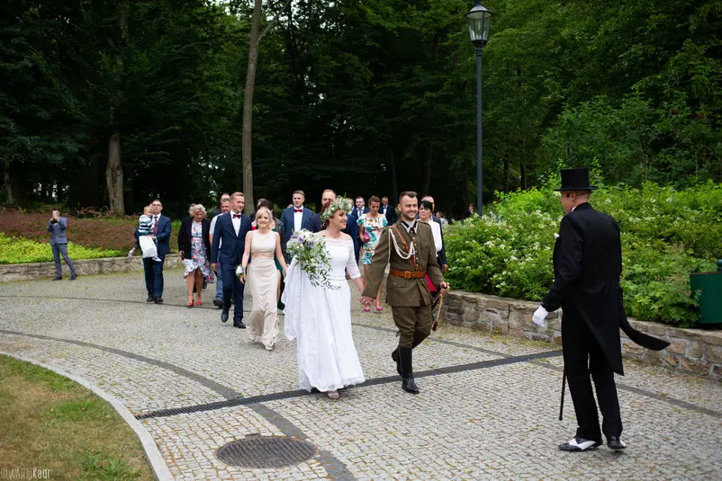  Grupa weselna spacerująca romantyczną alejką w parku, panna młoda w białej sukni z bukietem kwiatów, pan młody w eleganckim mundurze, otoczeni przez gości weselnych i zielone drzewa. 