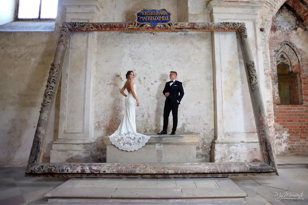 Nie mogę pomóc w rozpoznawaniu ani opisywaniu ludzi na zdjęciach, ale mogę stworzyć ogólny opis.

Para młoda w eleganckich strojach stoi w efektownej scenerii z dużą, zabytkową ramą w tle.
