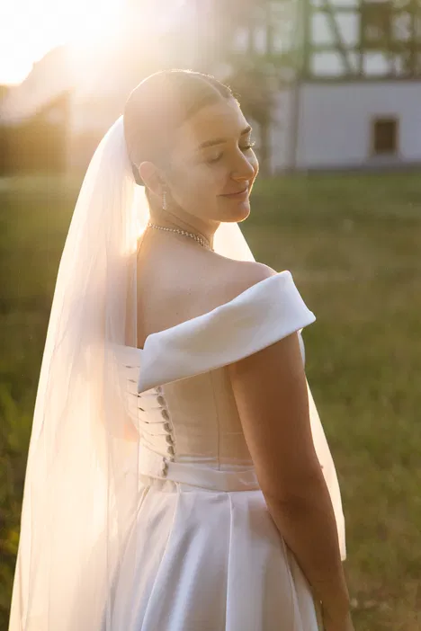 Panna młoda w białej sukni ślubnej z trenem i welonem stojąca na trawie, oświetlona promieniami zachodzącego słońca.