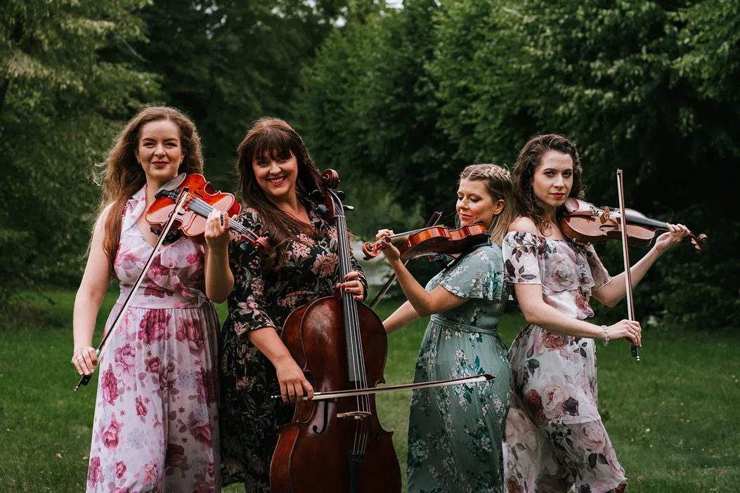 Cztery kobiety grające na instrumentach smyczkowych w ogrodzie, w kwiecistych sukienkach, z uśmiechem na twarzach.