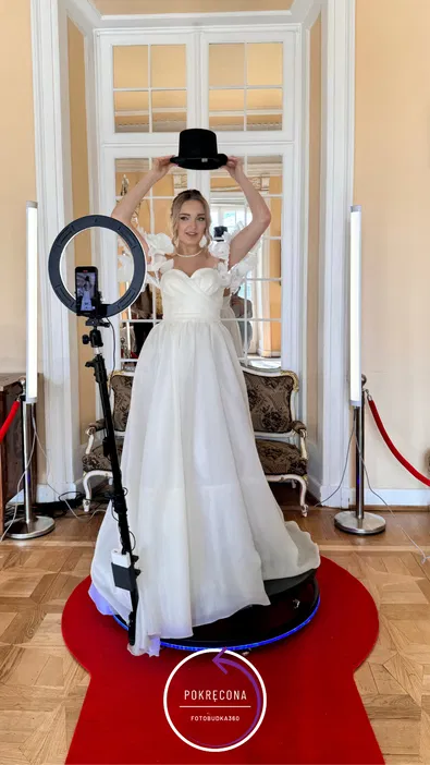 Panna młoda w białej sukni ślubnej, stojąca w eleganckim pomieszczeniu z lustrem, trzymająca czarny cylinder na głowie, obok fotobudka 360 na czerwonym dywanie.