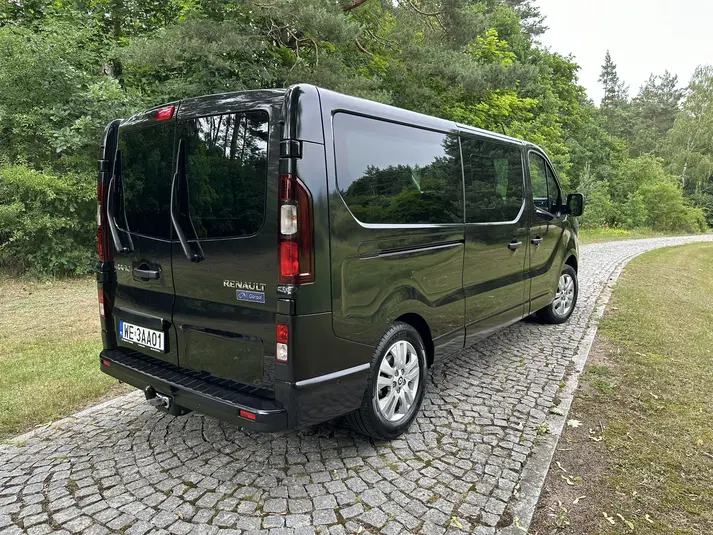  Czarny van Renault na brukowanej drodze otoczonej zielenią w leśnym otoczeniu. 