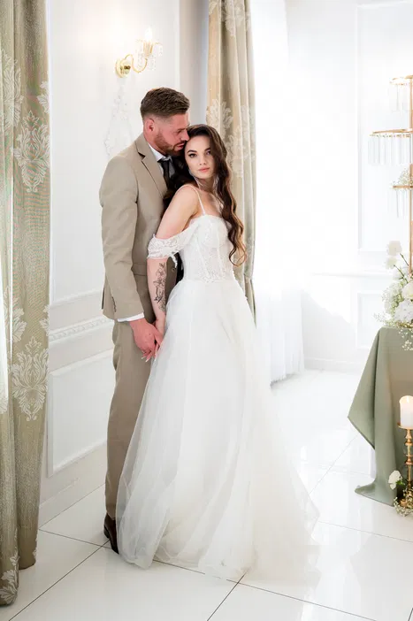 Para młoda w eleganckiej pozie na ślubie, panna młoda w koronkowej sukni ślubnej, pan młody w garniturze, romantyczne wnętrze z klasycznym wystrojem.