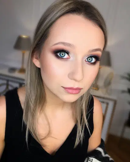  Młoda kobieta z blond włosami i intensywnym makijażem oczu, ubrana w czarny top, w eleganckim wnętrzu. 