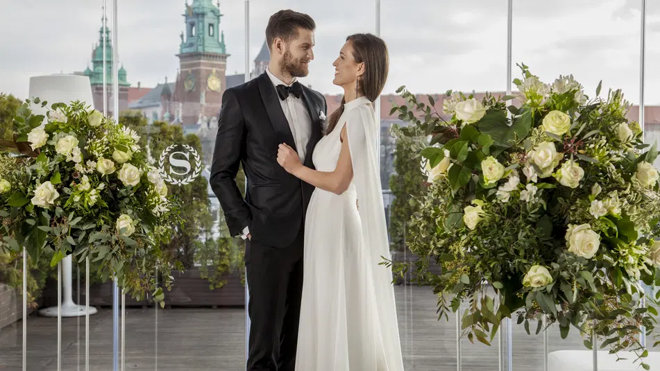  Para młoda w eleganckich strojach ślubnych na tle zamku, otoczona dekoracją kwiatową z białych róż. 