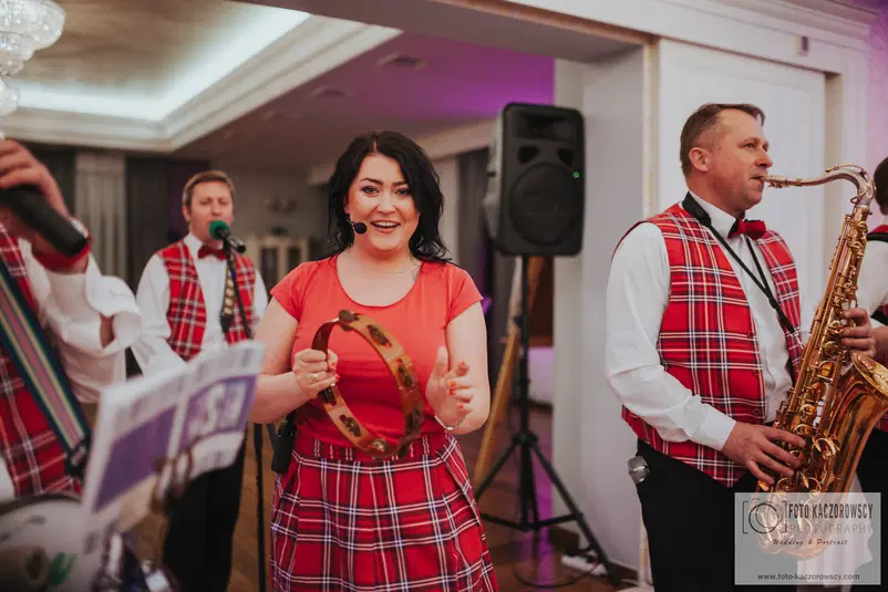  Zespół muzyczny występujący na scenie, kobieta z tamburynem, mężczyzna grający na saksofonie, tradycyjne stroje w kratę, wydarzenie muzyczne, koncert na żywo. 