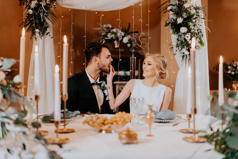  Romantyczna scena ślubna z parą młodą przy stole z dekoracją kwiatową i świecami, elegancka dekoracja w klimacie weselnym. 